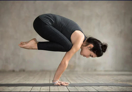 Woman in black workout clothes performing advanced crow yoga pose on wooden floor indoors