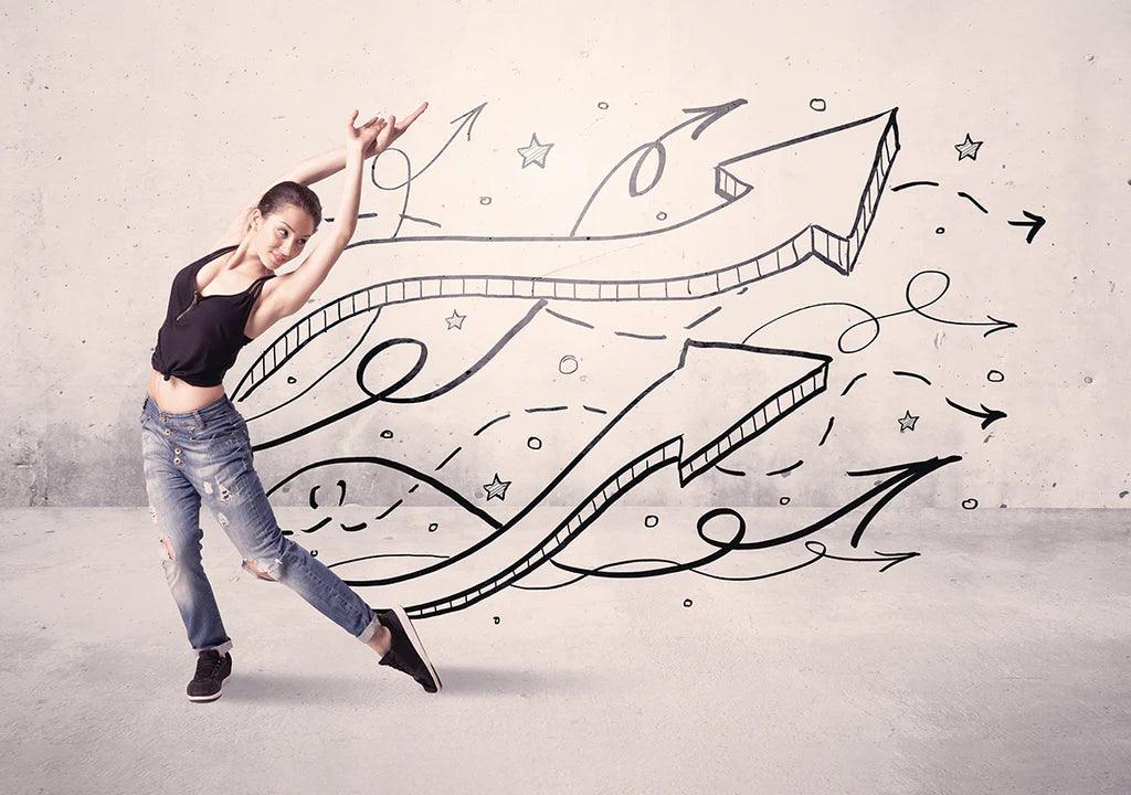 Young woman in casual outfit dancing next to wall with large hand-drawn arrows and sketches